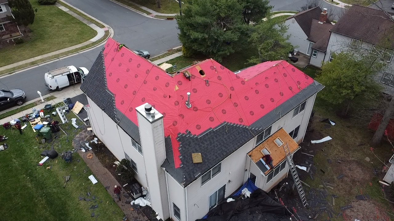 Aerial of a re-roof in progress, red underlayment down across the slopes with new shingles started along the lower courses