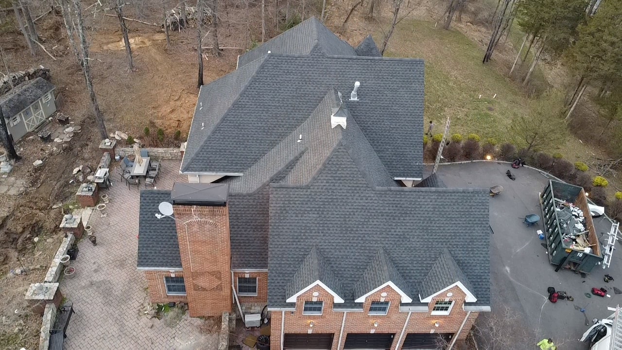 Aerial of a large brick home on a wooded lot with a finished dark gray shingle roof across all sections and dormers