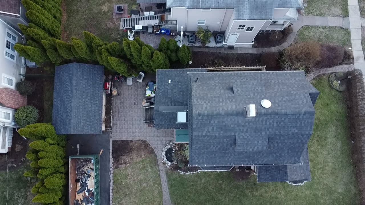 Aerial of a home with a finished dark gray shingle roof and a dumpster of torn-off roofing below