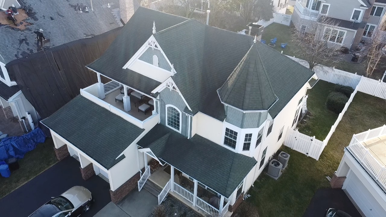 White home with a turret and wraparound porch, roof finished, a neighboring roof mid tear-off at the edge